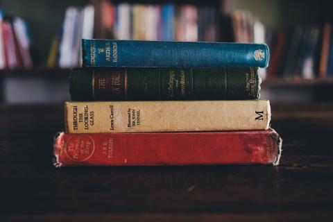 Four old books laying on their side stacked on top of each other on a table with a book shelf in the background.