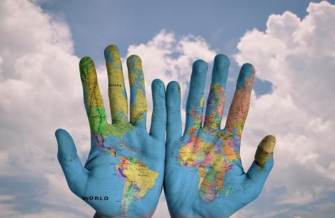 Two hands with palms painted with a map of the word in various colors in front of a blue sky with white, fluffy clouds.