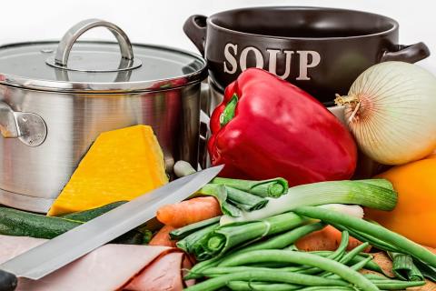 A silver cooking pan with vegetables and a knife in front of it with a black bowl with the word "soup" beside the pot.