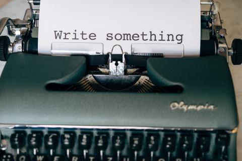 An old typewriter with a piece of paper in it with the words, "Write something" on the paper.