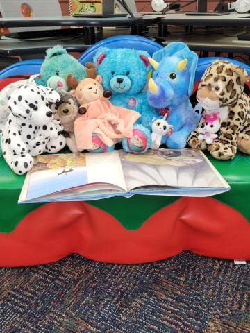 Stuffed animals (dog, hedgehog, bear, reindeer, bear, dinosaur, unicorn kitten, leopard and horse) sitting on a couch reading an open book.