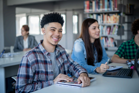 Smiling teens sitting at a table talking in a library with books and a laptop on the table.