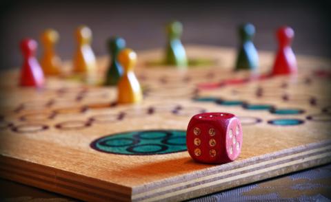 A wooden board game with a red die, two red game pieces, three yellow game pieces, two blue game pieces and one green game piece on the board.