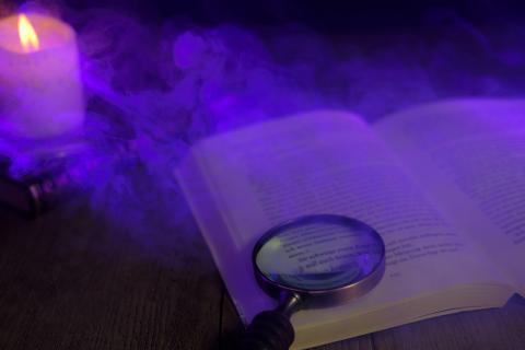 Image of an open book on a wooden table with a magnifying glass on top of it. There is also an lit candle on the table and fog swirling around.