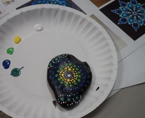 A white paper plate with a rock painted in a mandala pattern. There are different colors of paint also on the plate.