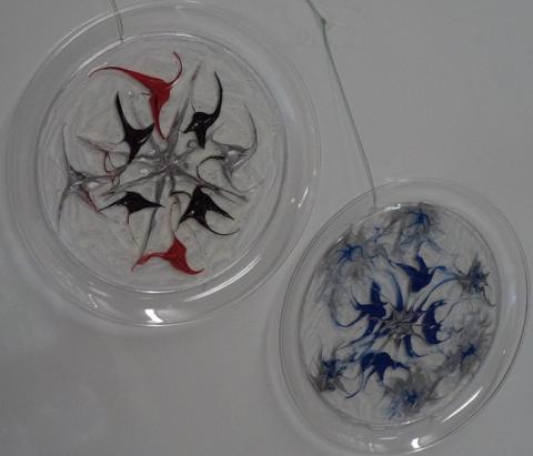 Two homemade suncatchers that are a painted design between two clear plates with a string hanging from the top of each.