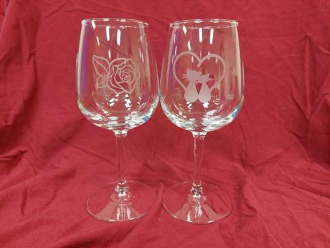 Two wine glasses with a rose etched on one and two cats in front of a heart etched on the other one. Both glasses are in front of a red backdrop.