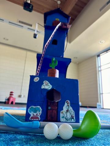 Image of a 4 story house built from cardboard/wood painted blue with Dr. Seuss characters on the front of it. There are two golf clubs and two golf balls in front of the house with a Cat in the Hat hat in the background.