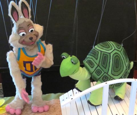 Image of a rabbit marionette wearing an orange and blue tank top standing next to a turtle marionette on a white bridge.