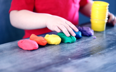 A small child's hand is playing with six piles of clay - red, orange, yellow, green, blue and purple. There are six small containers stacked next to the clay. The child is wearing a red shirt.