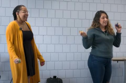 An African American woman wearing a black dress with a gold sweater standing next to a Hispanic woman wearing jeans and a gray sweater. Both have shaker eggs in each of their hands.