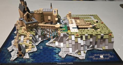 A LEGO model of Hogwarts Castle from the Harry Potter series is shown partial built. The castle has tan, gray and black bricks, featuring towers, turrets and connecting bridges. It sits atop rocky gray and green cliffs that rise from a base of blue LEGO bricks representing water. The model is mounted on a black base with a label that reads “Hogwarts™ Castle.”