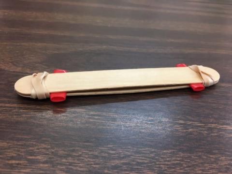 Image shows a simple homemade kazoo made from wooden popsicle sticks, rubber bands and pieces of red plastic straw. Two flat wooden sticks are stacked with a gap between them, held together at each end with beige rubber bands. Two short segments of red plastic straw are inserted between the sticks, one near each end. The object rests on a dark wood-grain tabletop.