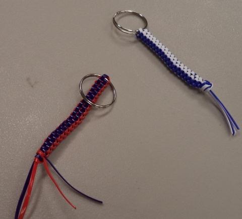 Image shows two handmade plastic lacing (rexlace) keychains on a gray surface. The keychain on the left uses red and dark blue plastic laces in a box stitch pattern, attached to a silver keyring, with the loose lace ends left hanging at the bottom. The keychain on the right uses white and dark blue laces in the same box stitch pattern, also attached to a silver keyring, with the loose ends left untied at the bottom.