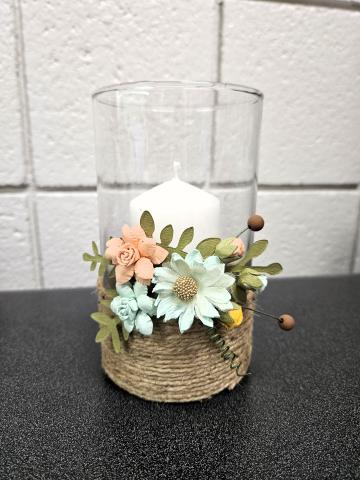 Image shows a decorative candle holder placed on a dark countertop against a white brick wall. The holder is a clear glass cylinder containing a white pillar candle. Around the base of the glass is a rustic embellishment: natural jute twine wraps around the bottom half, and an arrangement of artificial flowers and greenery is attached. The flowers include pale peach roses, light blue daisies with yellow centers and small blue rosebuds with green paper leaves and two small brown bead accents on wire stems.