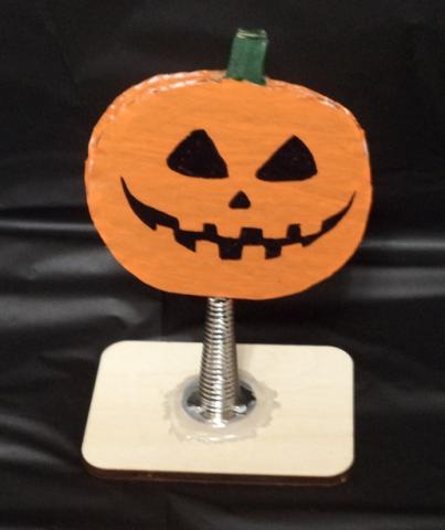 Image of a jack-o-lantern painted orange with a green stem and a black face. It is on top of a spring glued to a rectangular base.