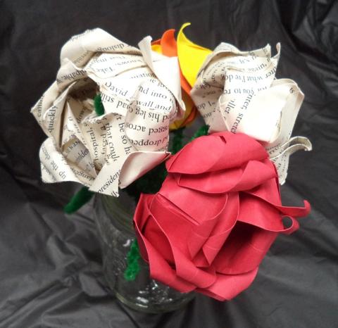 Image of a small bouquet of origami flowers arranged in a clear glass jar. The flowers are crafted from different types of paper: some petals are made from printed book pages with visible black text, while others are solid red or yellow. The stems appear to be made of green pipe cleaners. The background is a dark, crinkled fabric, which makes the flowers stand out.