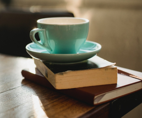 Two books on the corner of a wooden table with a green teacup and saucer on top.