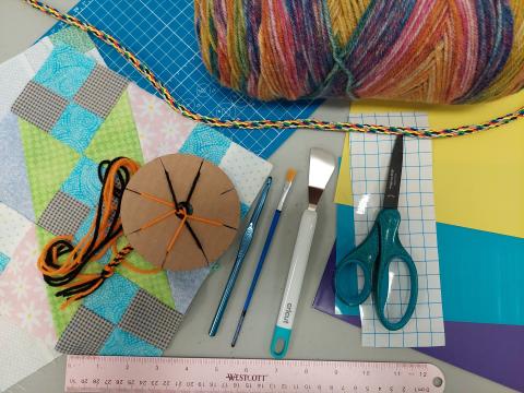Image of craft supplies with multi-colored yarn, scissors, paintbrush, crochet hook, fabric, contact paper, ruler, colored paper, Cricut spatula and a yarn wheel with some yarn on it.