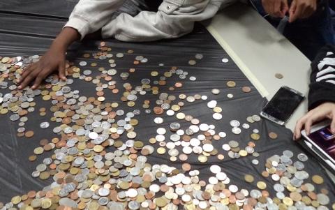 Image of 3 people looking at coins from different countries spread out on a black, plastic table cloth.