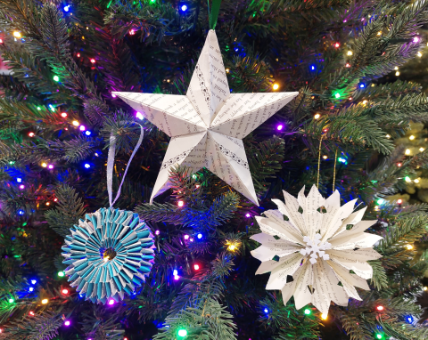 A woven wreath, a star and a snowflake disk ornament made out of book pages hanging on pine tree branches with colored lights around them.