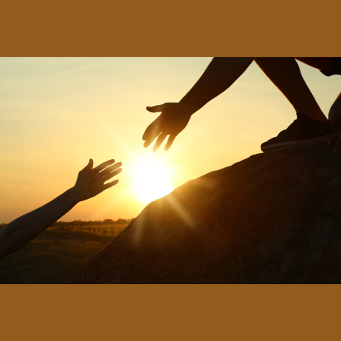 Image shows two silhouetted hands reaching toward each other at sunset. One person is standing or kneeling on top of a rock, extending a hand downward, while the other person reaches upward from below. The bright sun is centered between their hands, casting a warm golden glow across the sky. The background shows an open landscape with soft hills and a clear horizon. The scene conveys themes of help, connection and support.