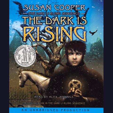 Cover of “The Dark Is Rising” by Susan Cooper that shows a snowy, wintry scene with a dark, leafless tree in the background. On the right side is a young boy with pale skin and dark hair. He looks directly at the viewer and holds a glowing circular symbol with a cross inside it. A white horse stands to the left of the boy, looking downward. Snow is falling throughout the scene, and the sky behind them transitions from deep blue at the top to a lighter blue near the horizon.