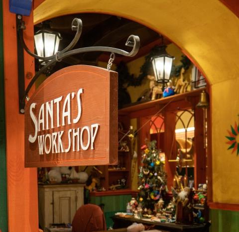 Image shows a festive indoor scene designed to look like Santa’s Workshop. In the foreground, a wooden hanging sign reads “Santa’s Workshop” in white letters. Behind the sign, the workshop is warmly lit with yellow and red walls, holiday decorations and a Christmas tree adorned with colorful ornaments. Toys, figurines and craft supplies are visible on tables and shelves, creating a cozy, cheerful holiday atmosphere.