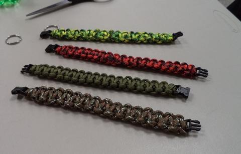 Image shows four handmade paracord bracelets laid out horizontally on a light-colored tabletop. Each bracelet has a black plastic buckle clasp. From top to bottom: bright green, yellow and black paracord; red and black paracord; solid olive-green paracord; and brown-and-green camouflage pattern. In the upper left corner of the image, there is a pair of scissors and a metal key ring resting on the table.