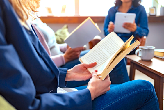 Women reading books and tablets around a coffee table with a cup of coffee on the table.